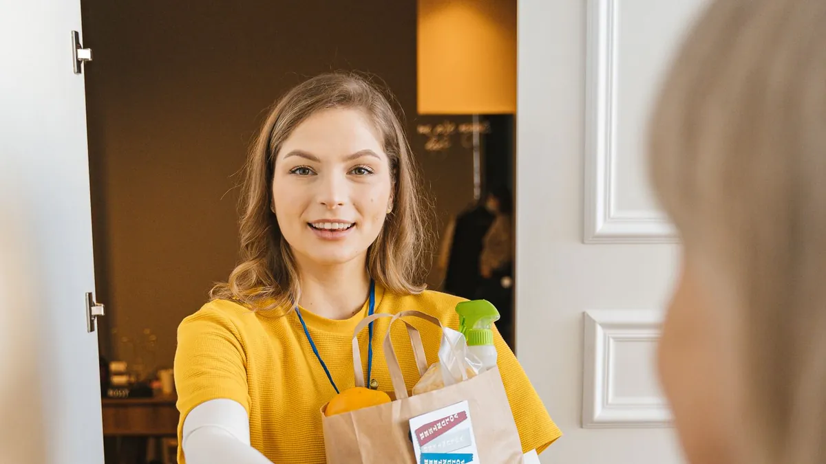Lächelnde Helferin bringt einer älteren Frau Einkäufe an die Tür