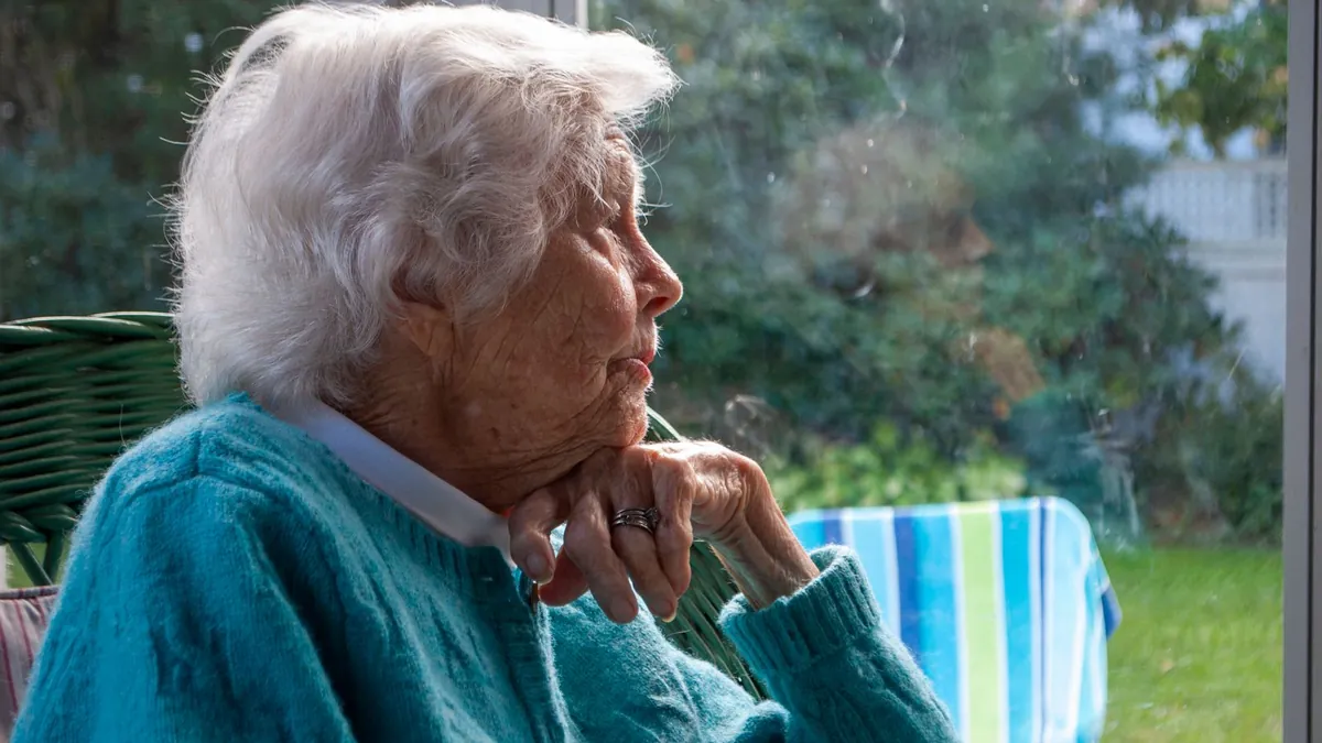 Ältere Frau sitzt nachdenklich am Fenster und schaut in den Garten