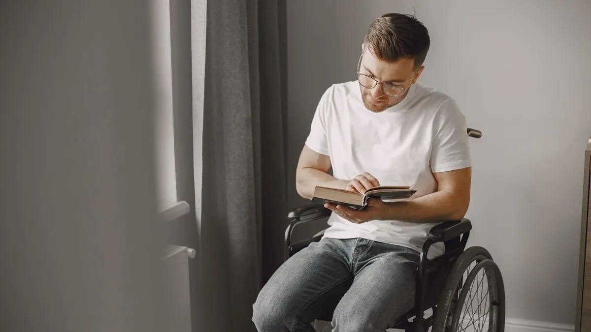 Mann im Rollstuhl liest zu Hause am Fenster ein Buch
